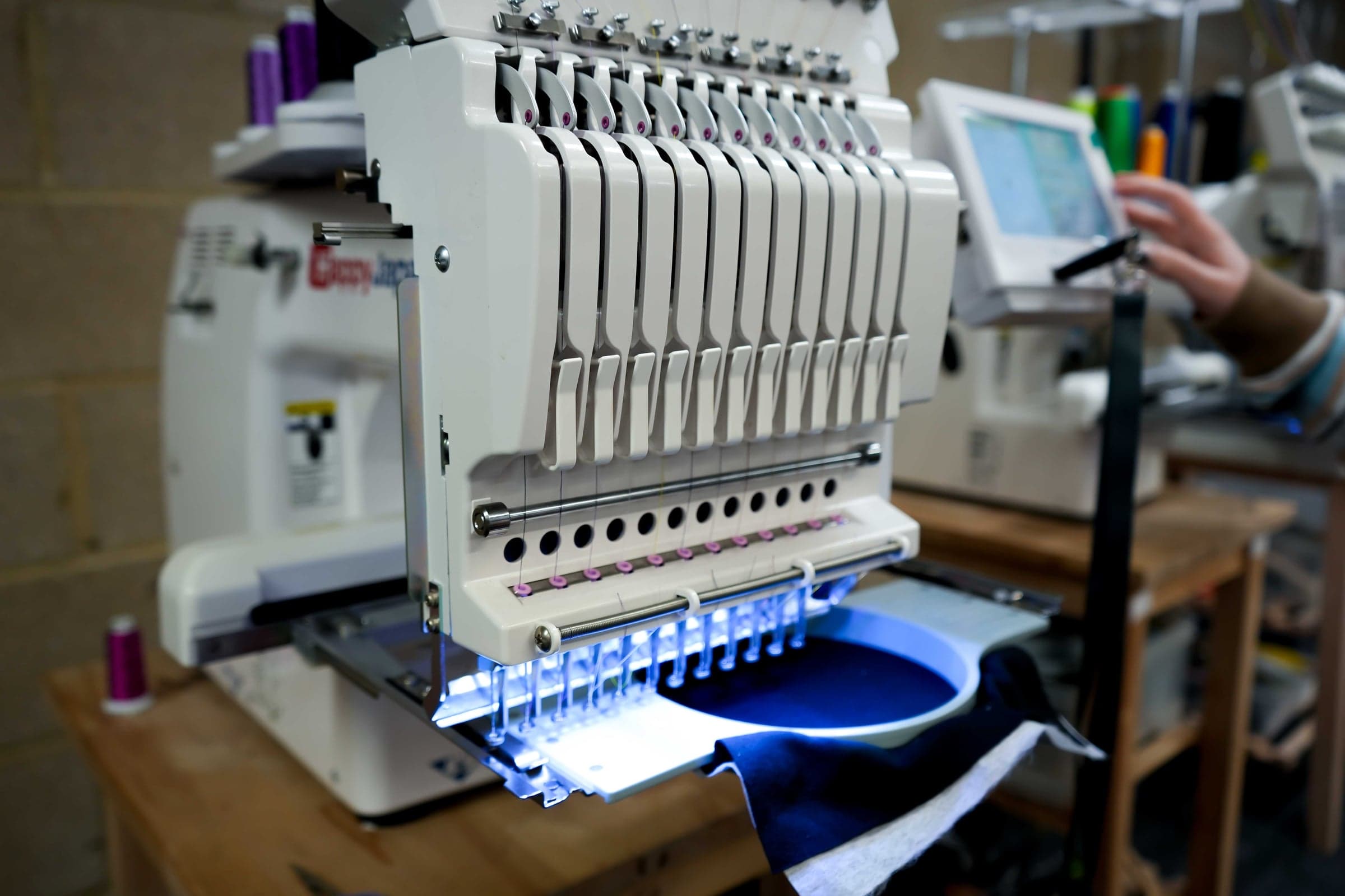 Multi-needle Happy Japan embroidery machine part-way through a run on navy fabric, with an operator setting the design on the touchscreen