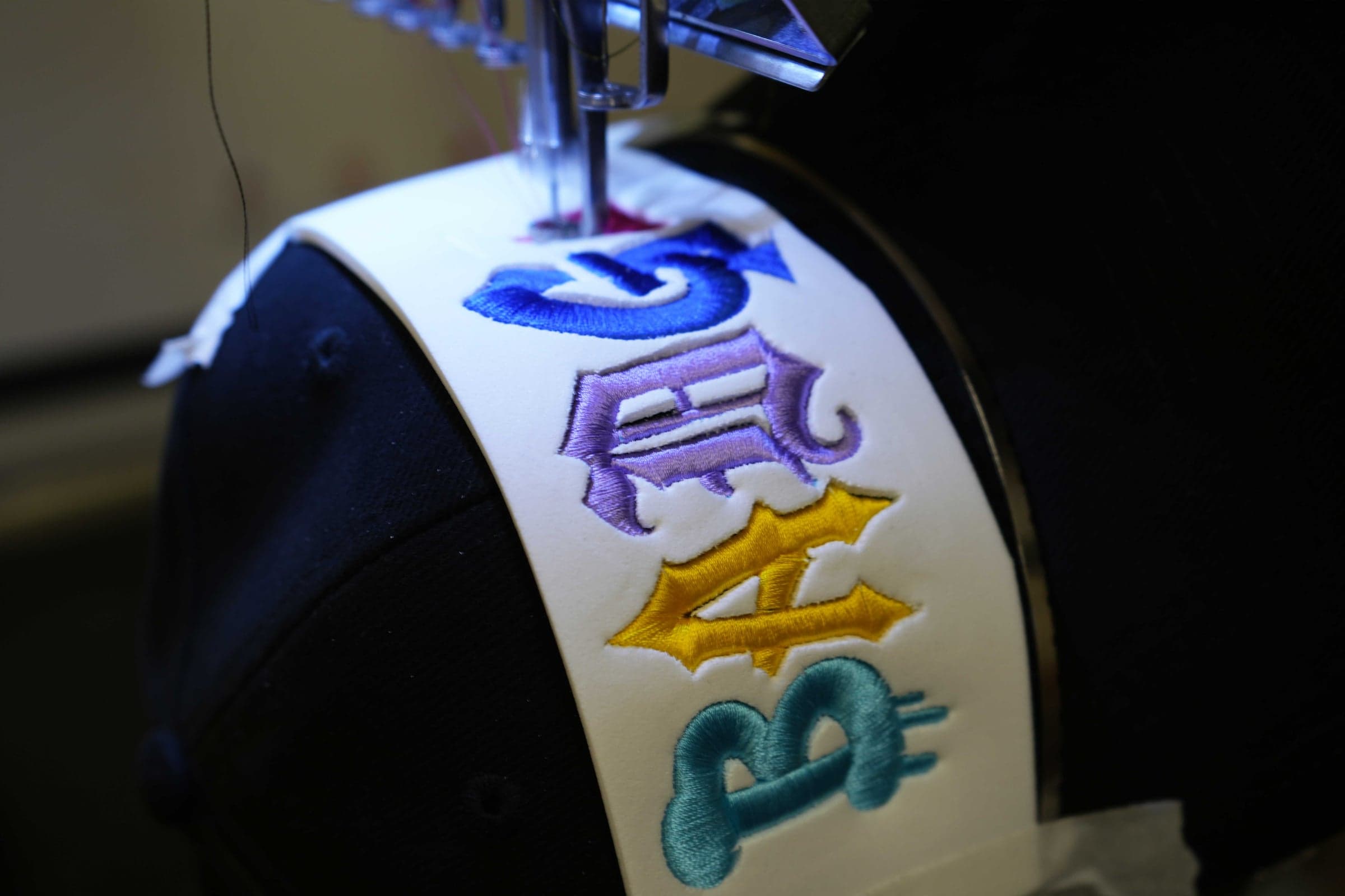 Close-up of a navy cap held in a cap frame, multi-coloured raised lettering stitched across the front panel beneath the needle head