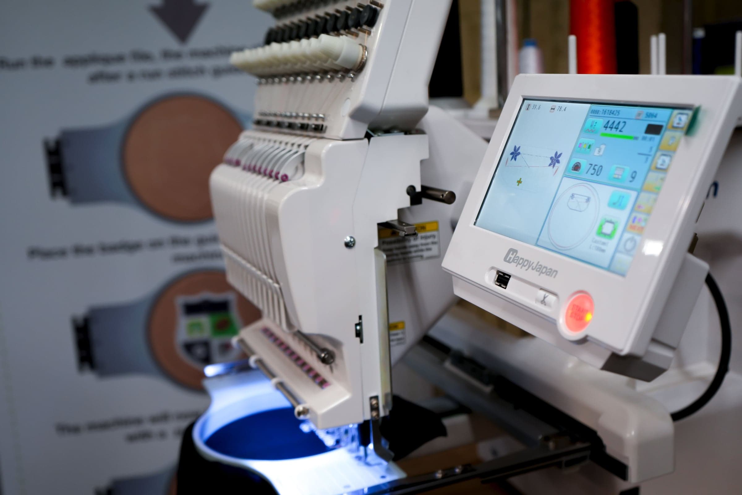 Happy Japan embroidery machine mid-run, its touchscreen showing a stitch count of 4442 at 750 rpm, in front of a badge placement instruction board