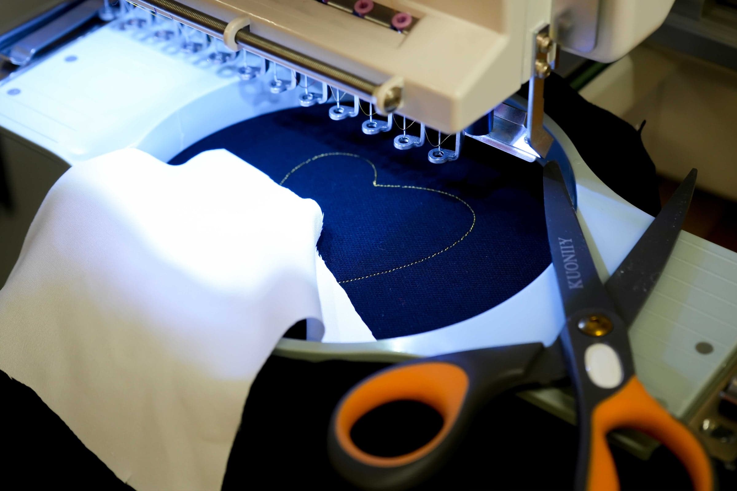 White applique fabric laid over navy material in an embroidery hoop, the tack-down outline already stitched and applique scissors ready to trim the excess