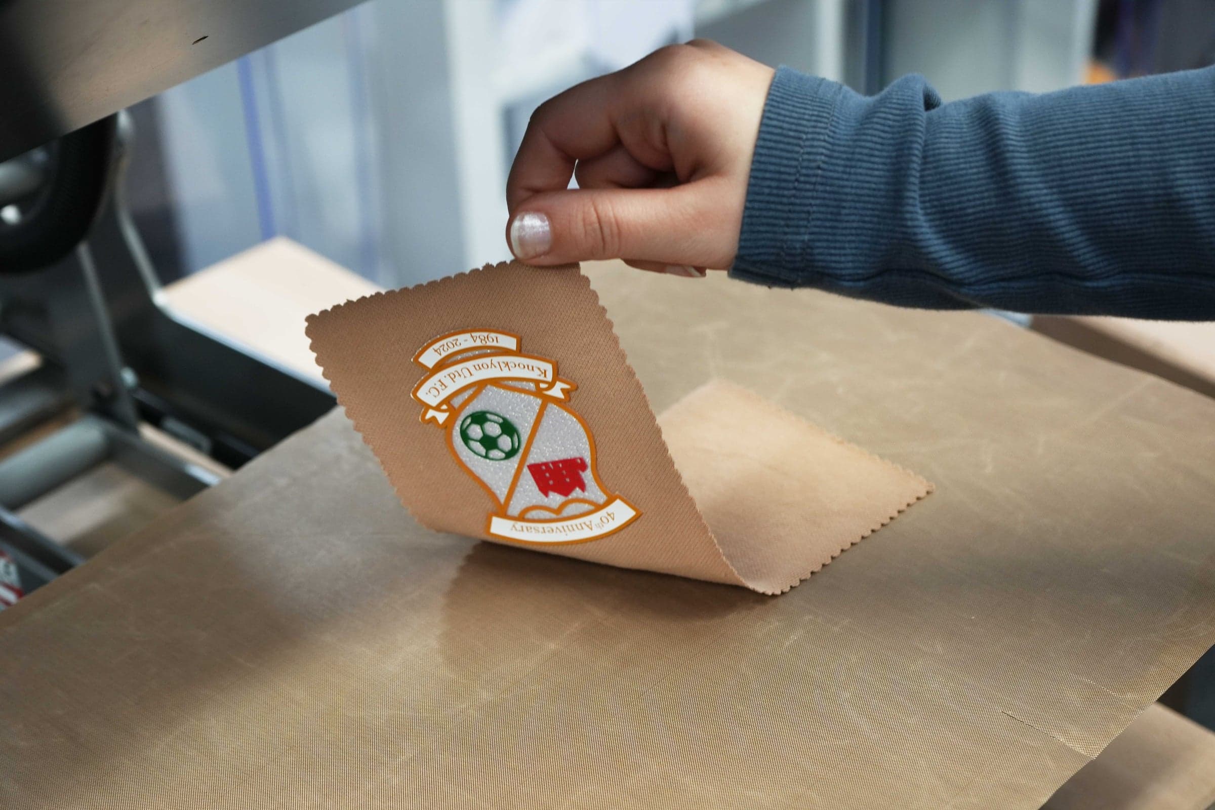 Hand peeling a custom DTF football-club crest transfer off the heat-press platen