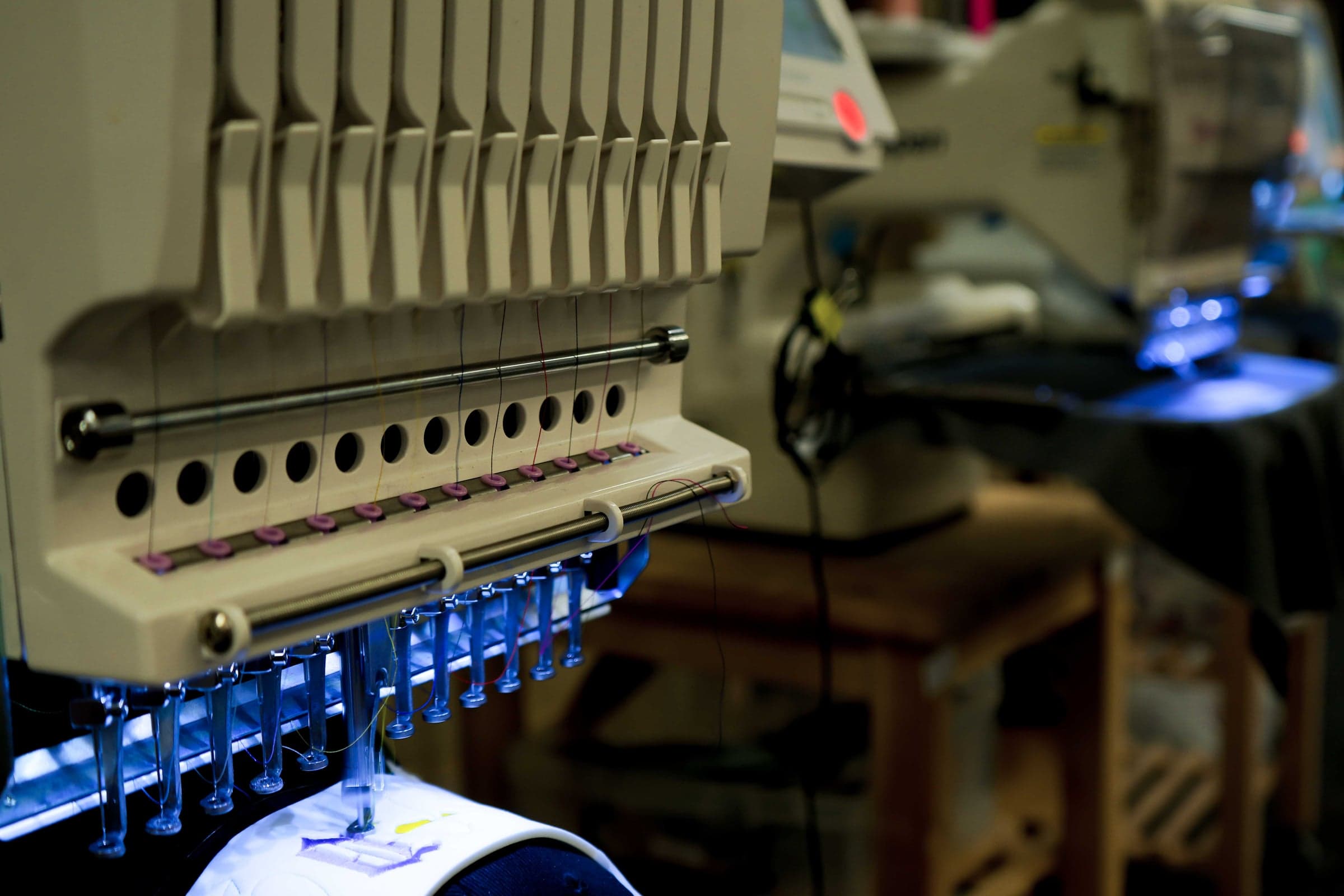Needle head of a multi-needle embroidery machine threaded with a full colour set, stitching a design held in the frame below